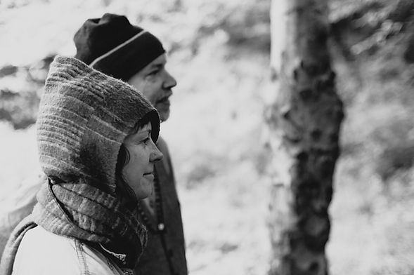 A black and white photograph of a man and a woman in outdoor clothing, stood side on to the camera, looking and smiling at something.