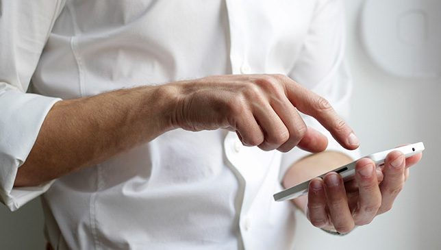 Un homme en chemise blanche utilise un smartphone. Il touche l'écran du téléphone. Fond neutre, ambiance concentrée.