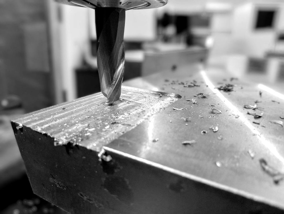 A milling machine cuts into a metal block, creating shavings. Black and white image, industrial setting, focus on the drill bit and surface.