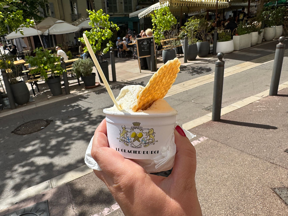 Glace à la main avec gaufrette sur une terrasse ensoleillée. Pot marqué "Le Glacier du Roi". Arrière-plan : cafés et verdure.