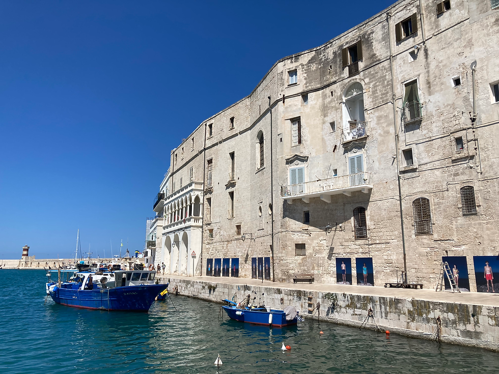 Bateau bleu amarré devant un vieux bâtiment en pierre, avec ciel bleu en arrière-plan. Des photos de personnes ornant le mur. Ambiance sereine.