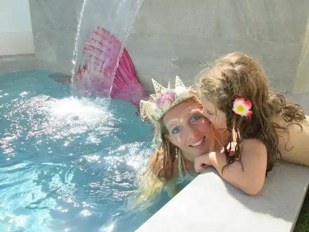 Femme déguisée en sirène, avec une couronne de coquillages, souriant dans une piscine aux côtés d’une petite fille avec une fleur dans les cheveux, sous un jet d’eau scintillant.