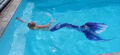 Une femme avec une queue de sirène de couleur bleu foncé nage en surface dans une piscine bleu clair.