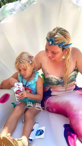 Femme en costume de sirène avec un diadème bleu, assise aux côtés d’une petite fille blonde en robe aquatique, découvrant un jouet dans un moment complice et joyeux.