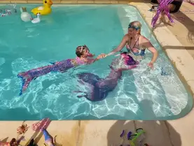 Petite fille portant une queue de sirène nageant dans une piscine cristalline, tandis qu’une femme sirène lui attrape la main, créant une scène féérique et complice.