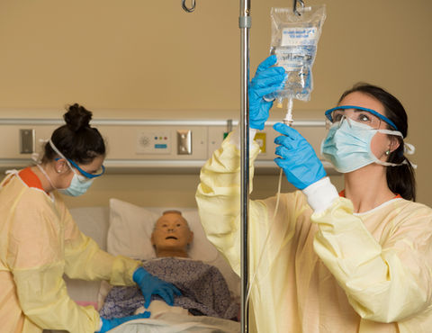 Nurses prepare an IV bag, medical simulation with patient in hospital bed.