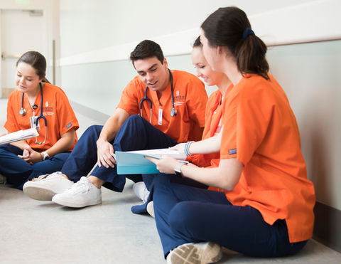 Four nursing students in orange shirts reviewing documents while sitting on floor.