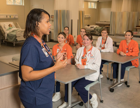 Nurse lectures students in a classroom setting, orange scrubs, AUCON.