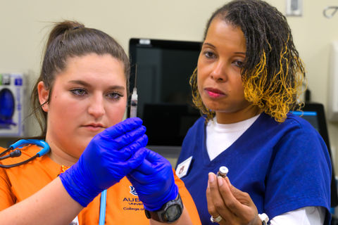 Two nurses practice with a syringe; clinical setting, AUCON College of Nursing.