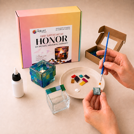 an array of art supplies set out for making a diy mosaic memorial luminary candle as part of the ugly art studio's grief support art kit. there are two hands visible, one holding a paint brush with glue and the other holding a mosaic tile with glue on it. there are tiles laying on a plate and a mosaic candle in process with blue and green tiles. 