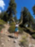 Runner in blue shorts and white cap joyfully raises arms on rocky forest trail. Clear blue sky; other hikers in background. Mammoth Trailfest