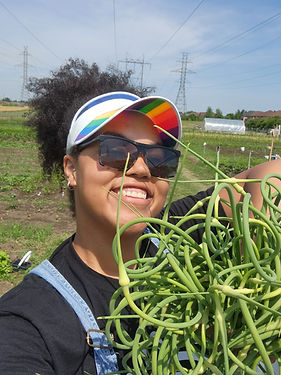 Claire holding a bunch of garlic scapes. She's wearing a rainbow visor, sunglasses, and a black shirt. Hydro towers and a hoop house are in the background.