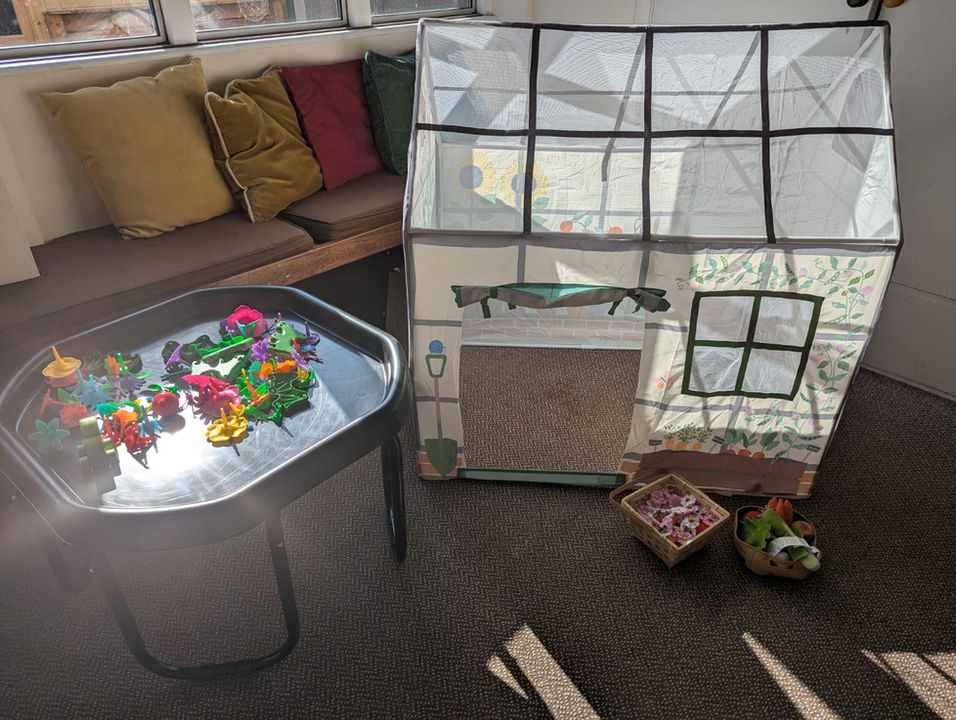 A tuff tray with flower toys next to a large play green house