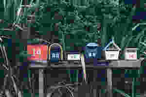 Row of 5 differently shaped mailboxes with the numbers 14, 16, 18, 20 and 28 on them. All perched on a wooden log with a backdrop of bushes.