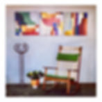 A green-cushioned Hans Olsen #532A teak rocking chair from Denmark, circa 1956 sits on a patio with three colorful abstract paintings on the wall. A potted plant and metal stand add decor.