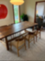 Wooden dining table with four Wegner dining chairs in a cozy room. A red abstract art piece and green plant are visible. Warm lighting and carpeted floor.