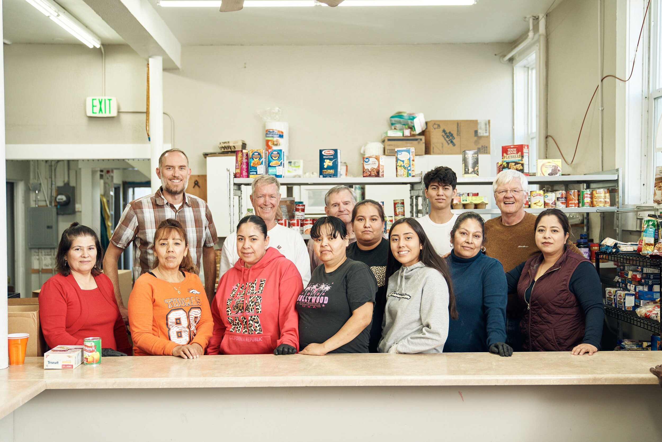 Twin Parishes Food Bank volunteer group beside shelves of donated food.