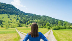 Woman in blue shirt stands at a forked path, facing green hills under a clear blue sky, conveying a sense of choice and exploration.