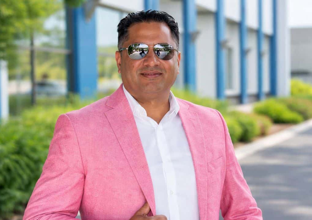 Smiling man in a pink blazer and sunglasses stands outdoors near a modern building and greenery.