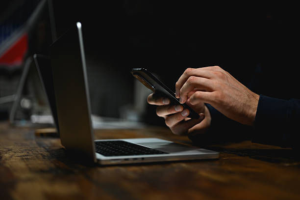 Hands hold a smartphone beside an open laptop on a wooden table. The setting is dimly lit, creating a focused and tech-oriented mood.