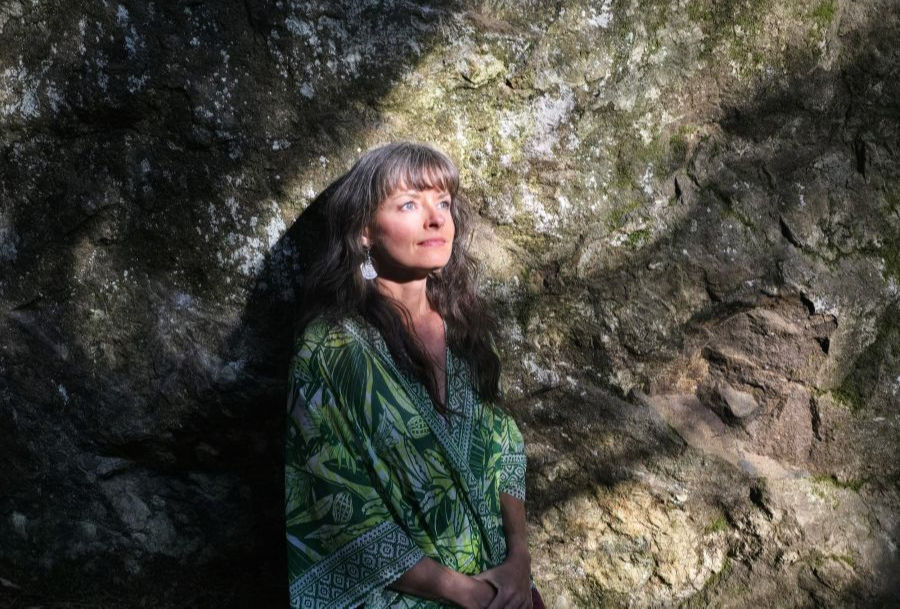 Woman in green patterned dress stands by a rocky wall, bathed in dappled sunlight, with a serene expression.