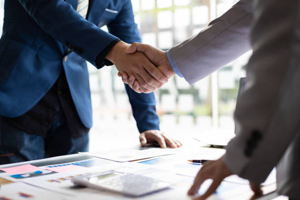 Business colleagues shake hands over charts in a bright office, suggesting a deal or agreement