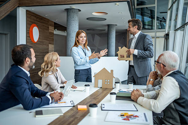 Business meeting with six people around a table. A woman gestures as a man holds a house model. Modern office, bright atmosphere.