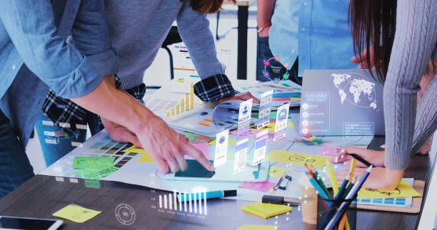 People collaborate at a table with colorful charts, sticky notes, and digital graphs, reflecting a tech-focused teamwork atmosphere.