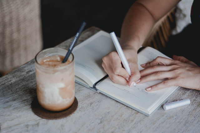 Hands write in an open notebook beside an iced coffee on a rustic table, creating a calm study scene.