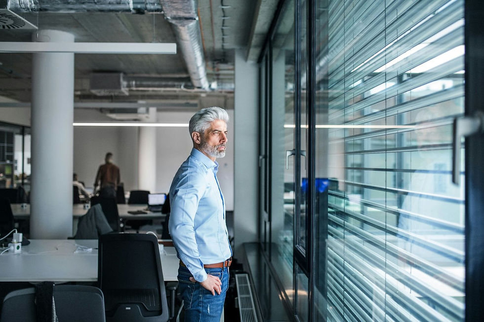 Man in blue shirt looks thoughtfully out of office window. Modern interior with desks, chair, and overhead ducts, creating a reflective mood.