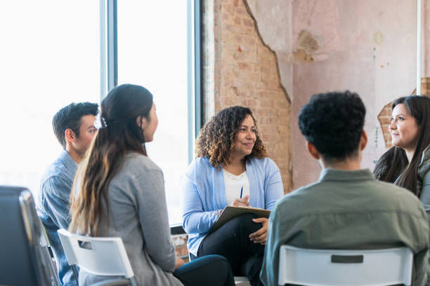 A diverse group of people sit in a circle, engaged in discussion, in a room with exposed brick walls and large windows.