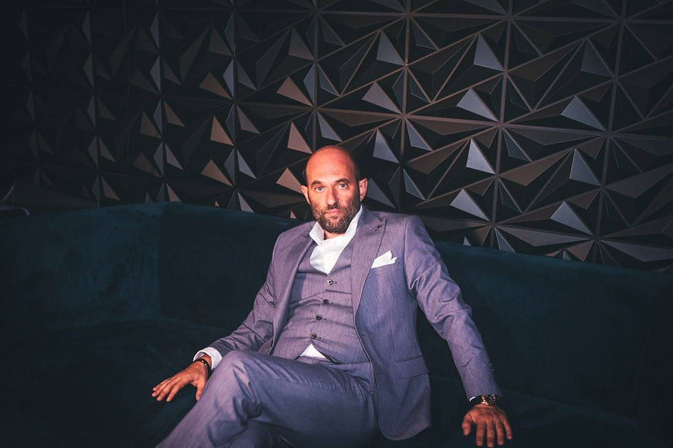 Man in a gray suit sits on a dark couch against a geometric-patterned wall. He appears relaxed, with a confident expression.
