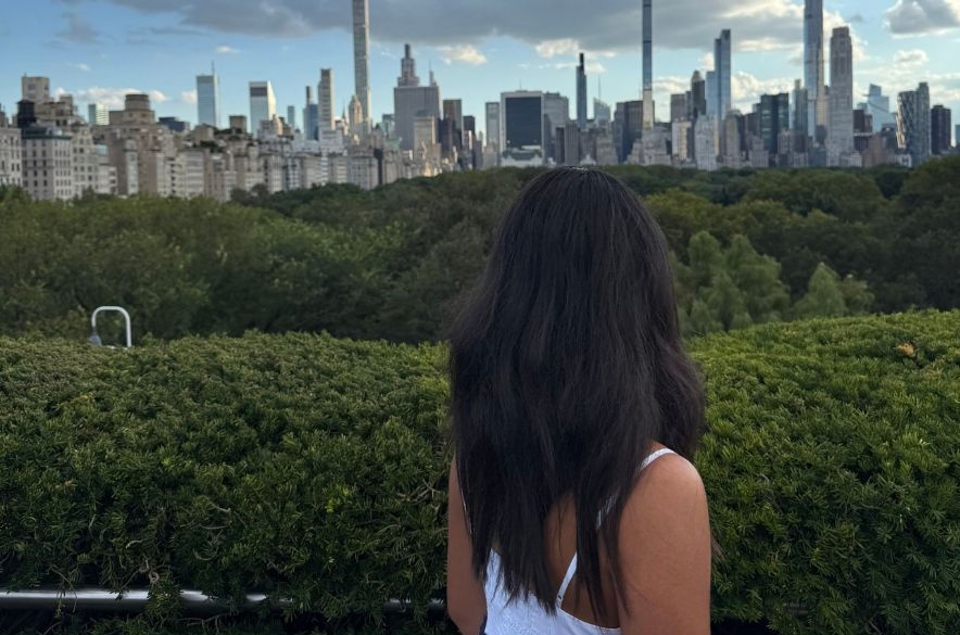 Woman in white top overlooks a city skyline with tall buildings under a cloudy blue sky. Green foliage in the foreground.