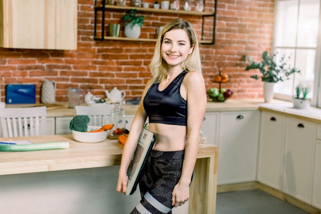 Smiling young woman in black workout clothes holds a tablet in a bright kitchen with brick walls and plants.
