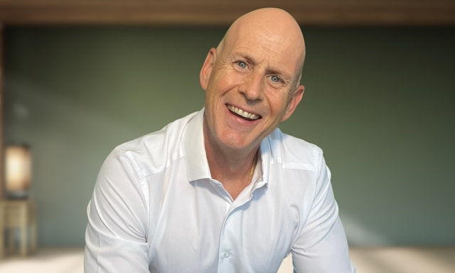 Bald man in a white shirt smiles warmly in a softly lit room with a blurred background. Calm and inviting atmosphere.