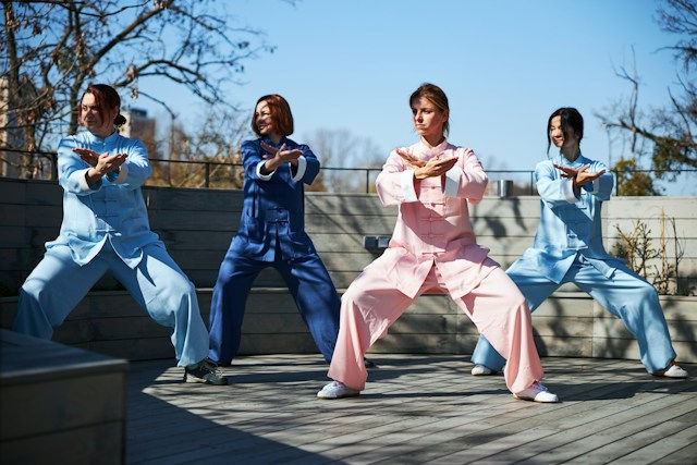 Four people in pastel martial arts outfits hold a wide stance pose on a sunny outdoor deck, with trees and blue sky behind them.