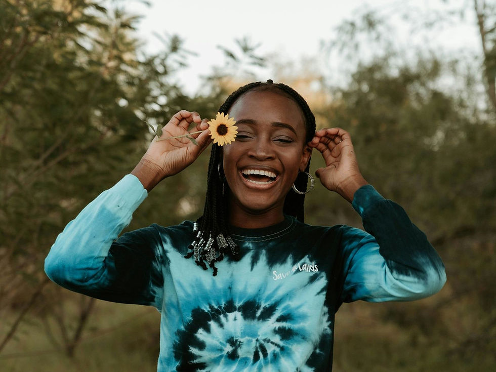 Woman smiling, holding a yellow flower near her eye. Wearing a blue tie-dye shirt in a lush, green outdoor setting. Joyful mood.
