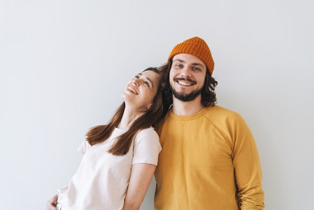 A smiling couple, a woman in a white shirt and a man in a yellow sweater and orange beanie, pose against a white background.