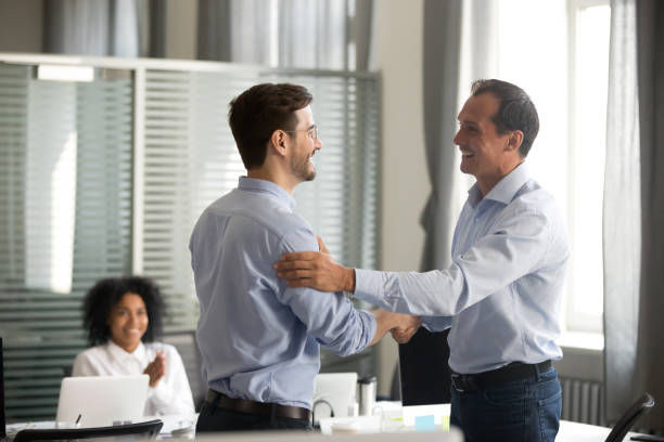 Two men shake hands and smile in a bright office, while a woman claps in the background.
