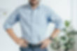 Man in a light blue shirt stands hands on hips in a bright white room beside a leafy plant, looking confident.