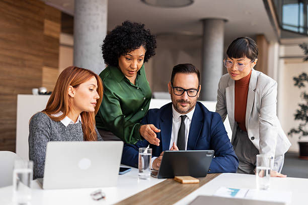 Four colleagues collaborate around a tablet in a modern office. A mix of attentive and focused expressions. Laptops and water glasses visible.