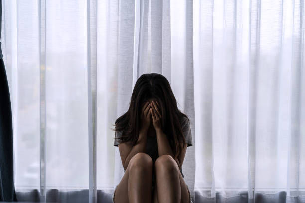 A person sits on the floor, head in hands, in front of sheer curtains. The mood is somber and reflective, with soft, muted lighting.