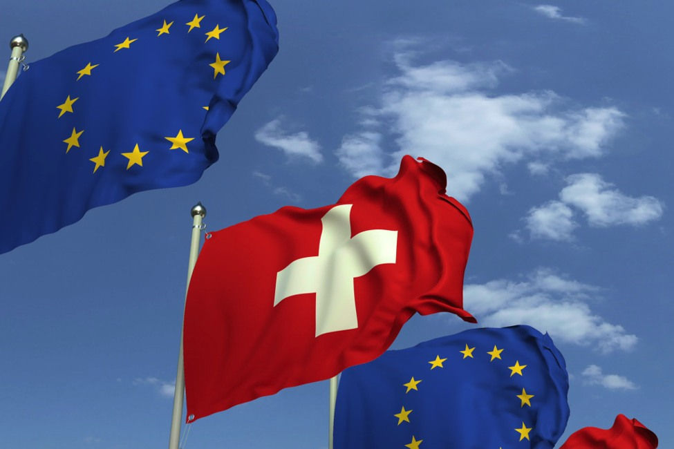 Swiss flag waves among EU flags against a blue sky with clouds.