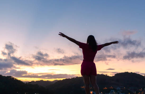 Silhouette of a woman in a red outfit with arms outstretched, facing sunset over hills. Sky has pink and blue hues, creating a peaceful mood.