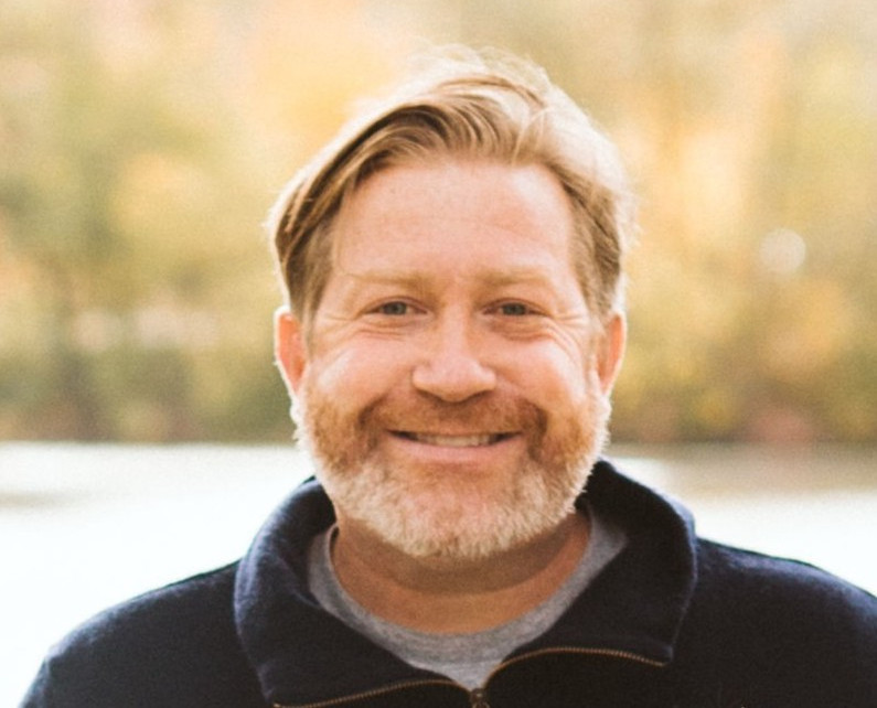 Smiling bearded man outdoors with a warm blurred autumn background, facing the camera.