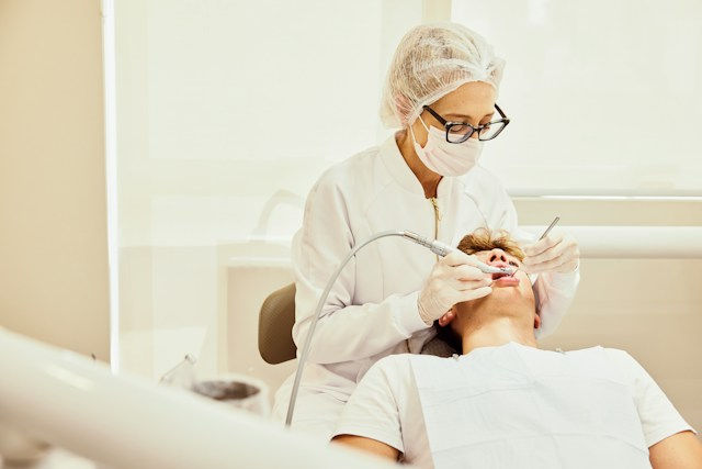 Dentist in mask and gloves works on reclining patient in a bright clinic, using tools during a dental exam.