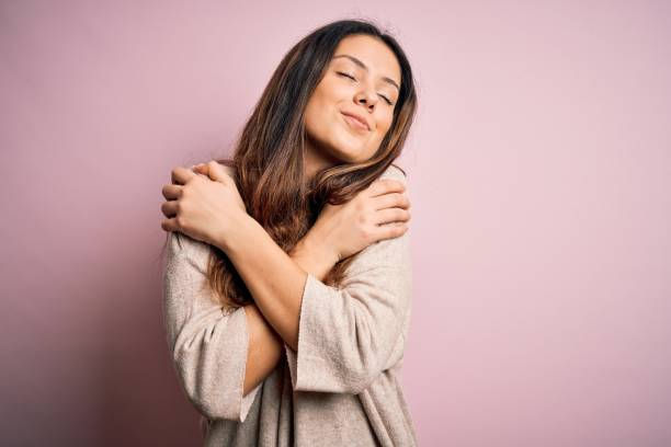 Woman wearing casual sweater standing over pink background hugging herself