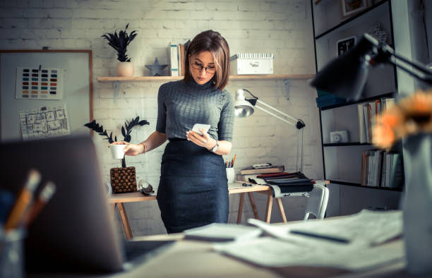 Woman in a gray sweater stands in an office, looking at her phone, holding a cup. Background has shelves, lamp, plants, creating a focused mood.