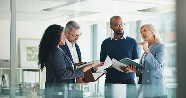 Four colleagues in a bright office discuss documents, one woman gesturing, with folders and a collaborative, focused mood.