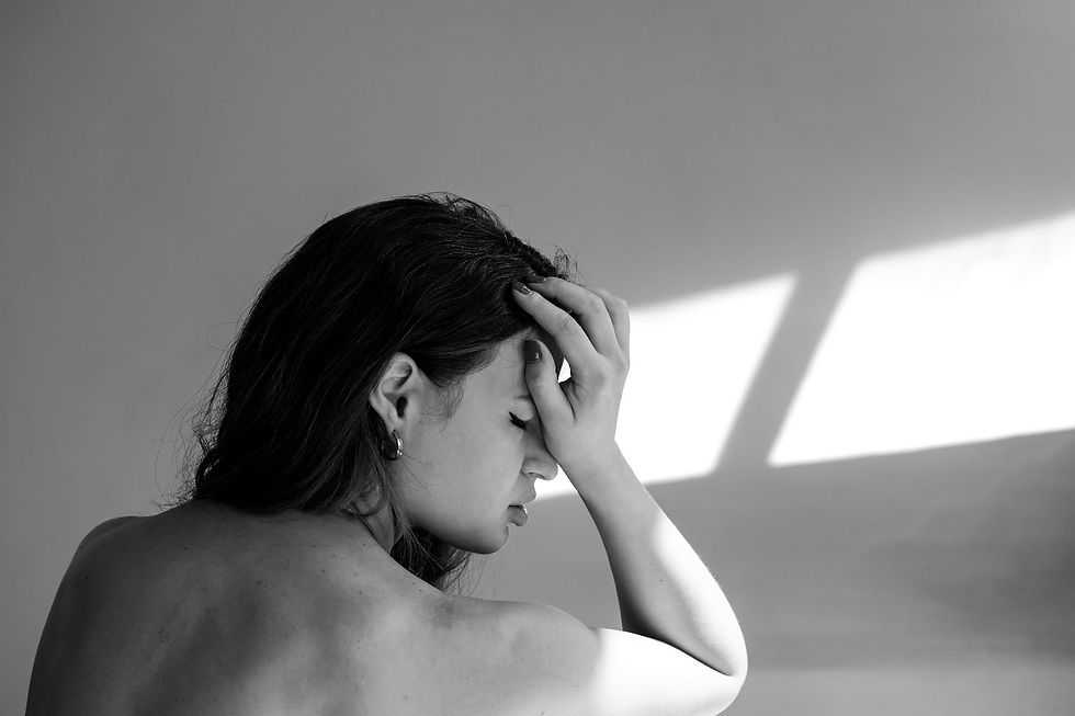 A woman holds her head in apparent distress, sitting topless in soft light against a plain background. The mood is contemplative and somber.